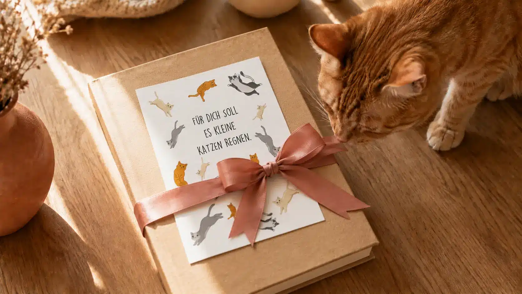 Katzenbuch als Geschenk verpackt mit rosa Schleife, handillustrierter Karte und Lesezeichen, dazu eine neugierige Katze