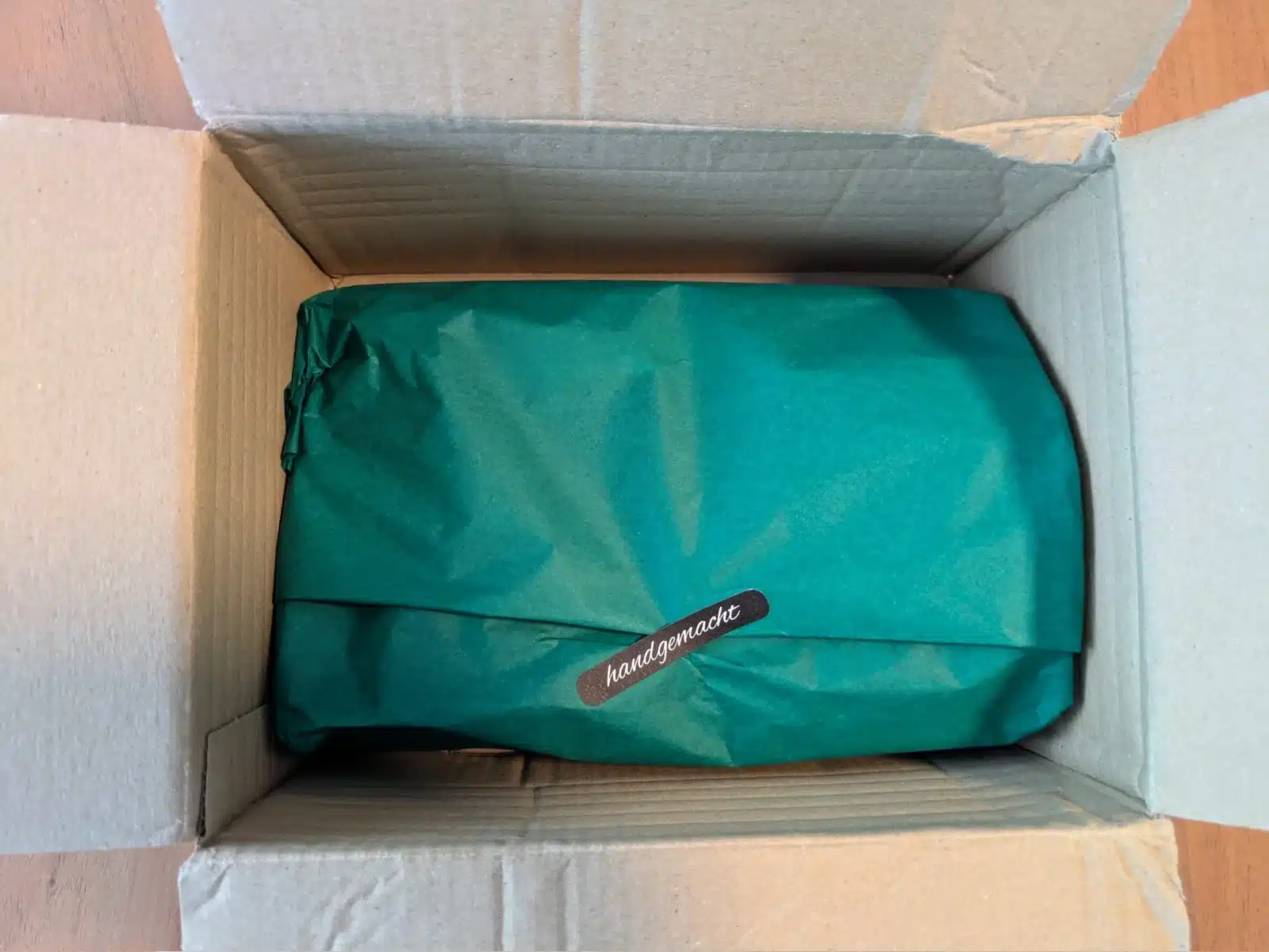 Geöffneter Versandkarton mit grünem Seidenpapier und einem Aufkleber mit der Aufschrift „handgemacht“.